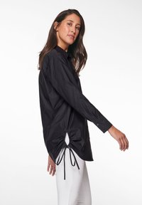 Schwarzes Langarmshirt aus leichtem Stoff, mit einem Kordelzugdetail an der Seite. Es hat einen kragenförmigen Ausschnitt und knöpfbare Manschetten.