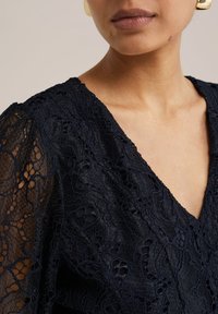Marinblå spetsblus med V-ringning och texturerade, intrikata mönster. Mjuk tyg med semi-transparenta, puffade ärmar och en delikat finish.