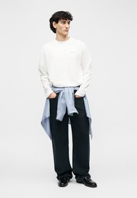 Jonge man met krullend haar die een witte trui, zwarte wijde broek, zwarte schoenen en een blauw gestreept overhemd om de taille gebonden draagt.