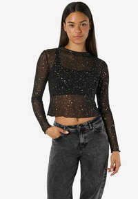 Črna prosojna dolgoletna majica z zlatimi pika, skrajšan dizajn in okroglo ovratnik, kombinirana z visokim pasom temno modrimi kavbojkami.