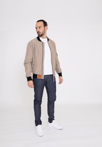 Beige bomberjack met zwarte accenten, ritssluiting en een oranje patch. Draagt over een witte shirt met donkere spijkerbroek en witte sneakers.