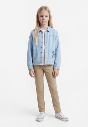 Jente står med et nøytralt uttrykk, iført en lyseblå denimjakke med broderi, hvit skjorte, beige bukser og hvite joggesko.