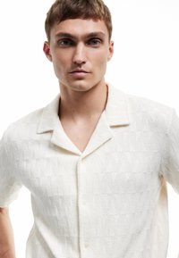 Jonge man met kort lichtbruin haar, gekleed in een witte textuur korte mouw knopshirt, kijkend recht vooruit tegen een witte achtergrond.