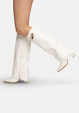 Leg-length view of a person wearing white knee-high boots with block heels and gold lock detail on the outer side.