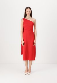 Robe rouge à une épaule en tissu lisse, longueur genou avec une silhouette cintrée et un drapé sur le côté. Portée avec des escarpins nude.