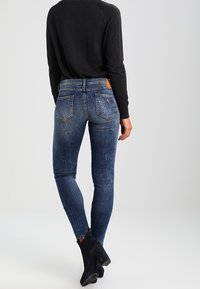 Jeans ajustados en denim azul desgastado con cintura de media altura, cinco bolsillos y una etiqueta de cuero marrón en la parte posterior. Combínalos con botas negras hasta el tobillo.