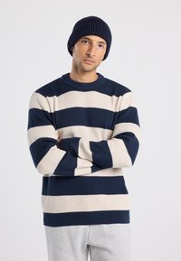 Uomo che indossa un berretto blu navy e un maglione a righe blu navy e beige con le braccia incrociate, in piedi contro uno sfondo chiaro e uniforme.