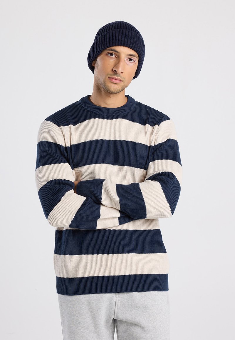 Uomo che indossa un berretto blu navy e un maglione a righe blu navy e beige con le braccia incrociate, in piedi contro uno sfondo chiaro e uniforme.