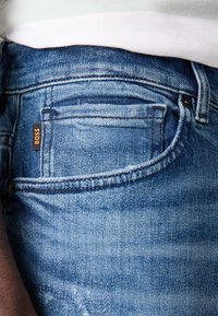 Kék denim farmernadrág, enyhén kopott mosással, elülső zsebbel és egy kis narancssárga "BOSS" címkével. A varrás jól látható a széleken.
