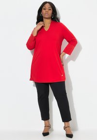 Rode tuniek met een V-hals en driekwartmouwen, gecombineerd met zwarte broek. Heeft een kleine gouden accent aan de zoom. Vloeiende textuur.