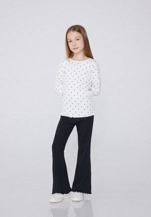 Haut blanc à manches longues avec des pois noirs, associé à un pantalon noir évasé. Encolure ronde et bords festonnés. Des chaussures blanches complètent la tenue.