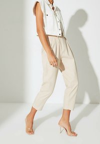 Femme portant un pantalon beige court, un débardeur blanc à boutons, et des sandales transparentes à talons hauts, se tenant avec un pied en avant.