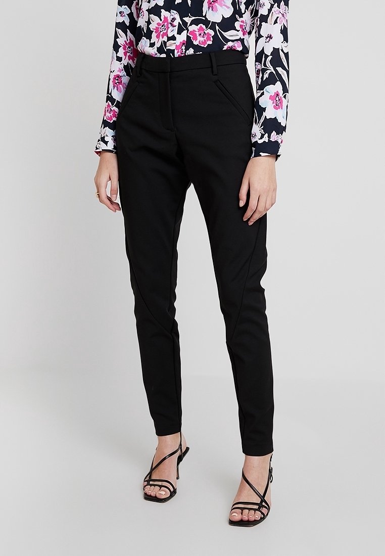 Femme portant un pantalon noir slim, un chemisier fleuri à manches longues et des sandales noires à talons avec brides, debout devant un fond uni.