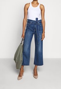 Spodnie dżinsowe o szerokich nogawkach z paskiem w talii, noszone z białym topem na ramiączkach i beżowymi obcasami, w połączeniu z zieloną torebką.