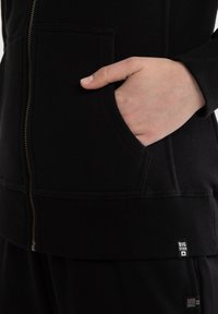 Ręka spoczywająca wewnątrz kieszeni czarnej bluzy z kapturem na zamek, z napisem "BIG STAR" w pobliżu krawędzi.