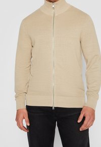 Beżowy prążkowany sweter na zamek z wysokim kołnierzem, noszony z czarnymi spodniami, widoczna przednia część tułowia i ramiona.