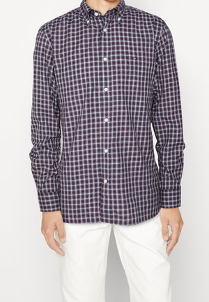 Homme portant une chemise à manches longues boutonnée avec un motif à carreaux bleu marine, blanc et bordeaux et un pantalon blanc sur un fond uni.