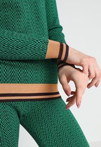 Grünes gestricktes Outfit mit einem Fischgrätenmuster. Die Bündchen sind orange mit braunen Streifen, die zum Taillenbund passen. Das Material wirkt weich und elastisch.