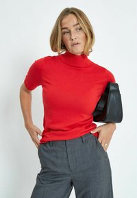 Suéter rojo de manga corta y cuello alto en una suave tela de punto, combinado con pantalones grises de corte ajustado y un bolso negro.