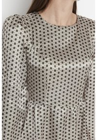 Femme portant une robe en satin à manches longues avec un motif carreaux noir et blanc, un col rond et une taille cintrée.