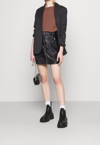 Blazer noir à rayures sur un top marron, associé à une jupe en cuir noir avec une fermeture éclair et des poches. Bottines noires et petit sac.