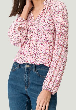 Vrouw draagt een witte blouse met lange mouwen met een klein roze bloemenprintje en blauwe jeans met hoge taille, hand rustend nabij de nek.