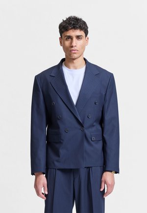 Giovane uomo rivolto in avanti che indossa una giacca doppiopetto a righe blu navy sopra una maglietta bianca.