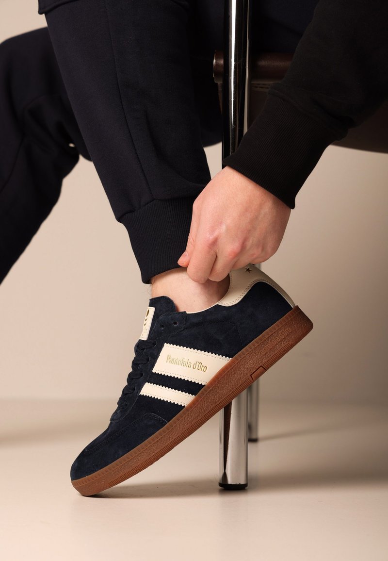 Granatowe zamszowe sneakersy z białymi paskami, kremowymi akcentami i gumową podeszwą. Jedna noga jest regulowana, odsłaniając teksturowaną podeszwę.