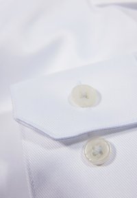 Witte overhemdkraag met zichtbare stiksels en twee lichtgekleurde knopen, voorzien van een subtiele gestructureerde patroon.
