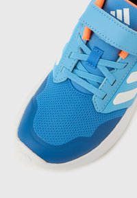Chaussure de sport bleue avec une tige en mesh respirant, des superpositions texturées, une sangle réglable et des accents blancs sur les côtés et l'avant.