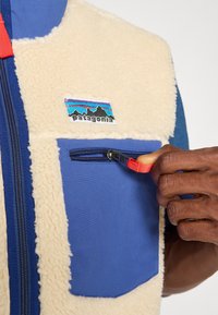 Fleece vest with cream and blue panels, front zip closure, and a zip pocket; features Patagonia logo patch on the chest.