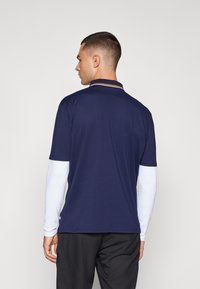 Ναυτικό πόλο μπλουζάκι με γιακά διαθέτει λευκά μακριά μανίκια. Ο γιακάς έχει πορτοκαλί και λευκές ρίγες. Το υλικό φαίνεται ελαφρύ και απαλό.