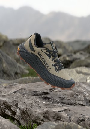 Chaussure de randonnée Merrell beige et noire avec une semelle marron flottant au-dessus d'un terrain rocheux et d'un arrière-plan montagneux flou.