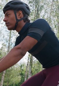 Maillot de cyclisme noir à manches courtes avec panneaux texturés et shorts bordeaux ; cycliste portant un casque noir devant un arrière-plan flou de feuillage.