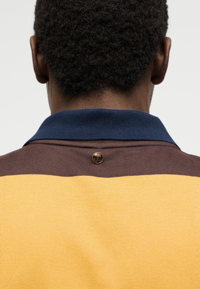 Άτομο που φαίνεται από πίσω φοράει κίτρινο και καφέ πουκάμισο με μπλε γιακά και ένα μικρό στρογγυλό κουμπί στο λαιμό.