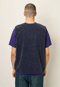 Tmavomodrý fleecový vest s textúrovaným, mäkkým povrchom, širokými výstrihmi na ruky a voľným strihom, nosený cez fialové tričko.