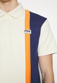 T-shirt polo crème avec des rayures verticales bleu marine et orange et un petit patch logo FILA sur la poitrine, porté par une personne avec un tatouage sur l'avant-bras.