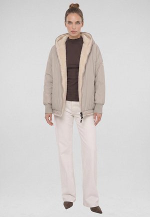 Vrouw staande, gekleed in een beige capuchonjas met bontvoering, donkerbruine top, witte broek en bruine puntige schoenen tegen een effen lichte achtergrond.