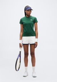 Γυναίκα με πράσινο πουκάμισο και λευκά σορτς που κρατά ρακέτα τένις, φοράει μπρασελέ, προβοσκίδα και λευκά αθλητικά παπούτσια, στέκεται σε λευκό φόντο.