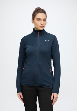 Femme portant une veste zippée bleu foncé avec des tirettes de fermeture éclair orange et un petit logo blanc sur la poitrine, debout devant un fond uni.