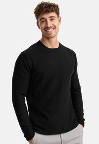 Zwarte langwerpige crewneck shirt met een zachte textuur en een relaxte pasvorm, gecombineerd met lichtgrijze broek, voor een witte achtergrond.
