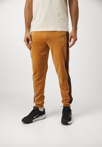 Oranžne velur joggers hlače z modrimi stranskimi črtami in majhnim logotipom, z elastičnimi manšetami in pasom na vrvico. Črne športne copate.