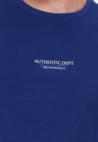 Gros plan d’un t-shirt bleu à col rond avec un texte blanc et bleu clair indiquant Authentic Dept Studio Collection Pier One Originals.