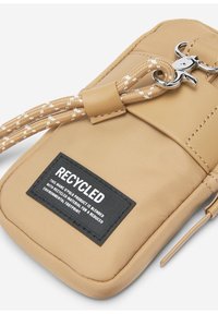 Custodia per telefono riciclata in tonalità marrone chiaro con una finitura testurizzata, caratterizzata da un'etichetta nera prominente, una tracolla intrecciata beige e dettagli in metallo argento.