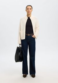 Cárdigan de punto crema con cuello chal, combinado con una blusa negra y vaqueros de pierna ancha en azul oscuro. Como accesorio, un bolso tote negro.