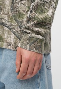 Μπλουζάκι με μακριά μανίκια σε πράσινο και καφέ παραλλαγή, υφασμάτινη υφή, με ριγωτά μανίκια, συνδυασμένο με ανοιχτού μπλε τζιν παντελόνι.