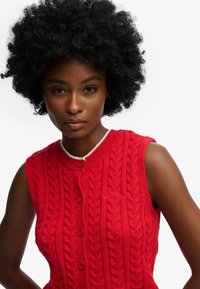 Pull sans manches rouge en tricot avec un motif torsadé, doté d'un col rond, d'une bordure blanche et d'une fermeture à boutons à l'avant.