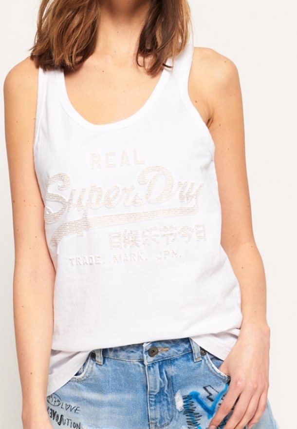 Femme portant un débardeur blanc SuperDry et un short en jean bleu avec un texte graphique, debout, une main dans la poche.