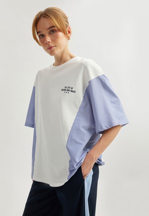 T-shirt en coton blanc avec des manches surdimensionnées rayées bleu et blanc. Présente un texte brodé : "LA VILLE DES RÊVES ET DE LA MAGIE." Coupe décontractée.