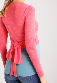 Růžový top s dlouhým rukávem a designem na zavázání vzadu, vrstvený přes světle modré tílko, v kombinaci s tmavými džíny. Měkká textura a volný střih.